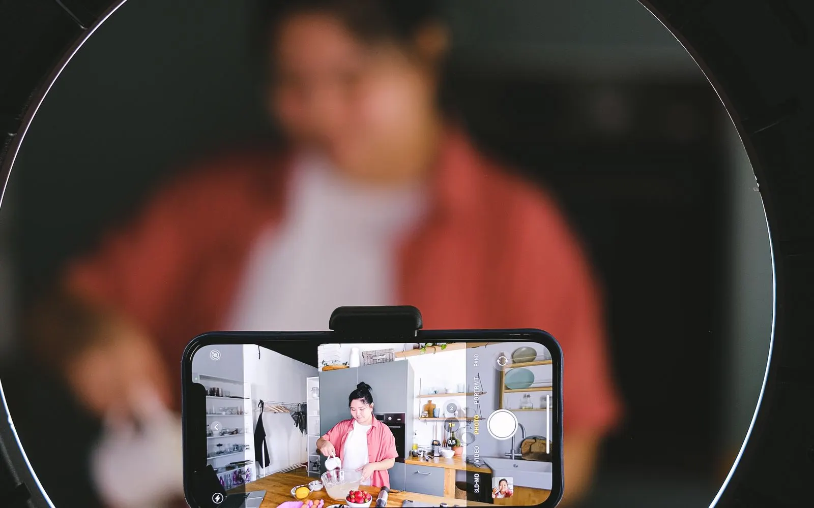 A smartphone on a ring-light stand filming a creator at work in a kitchen.