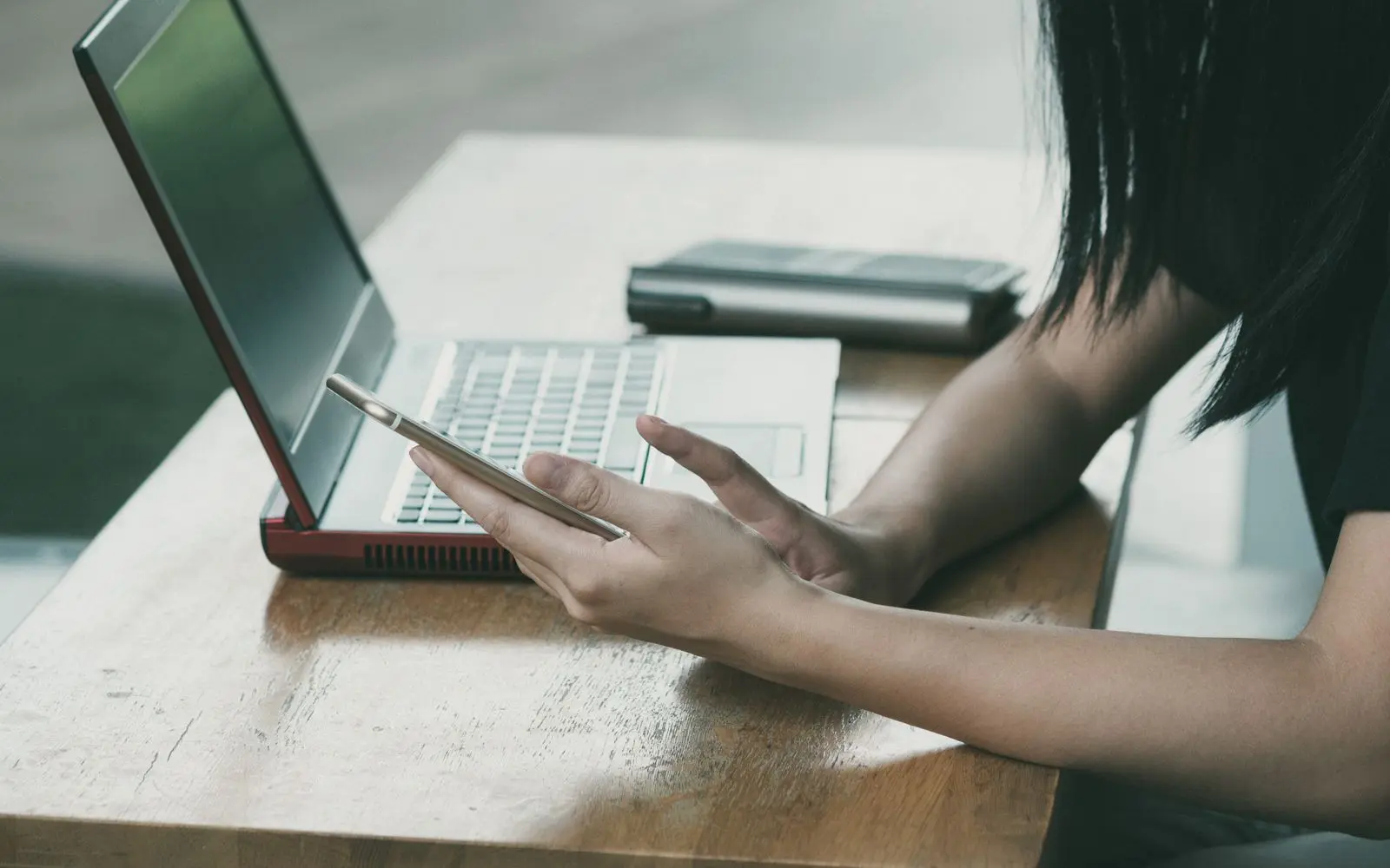 A person at a wooden desk using a phone beside an open laptop.