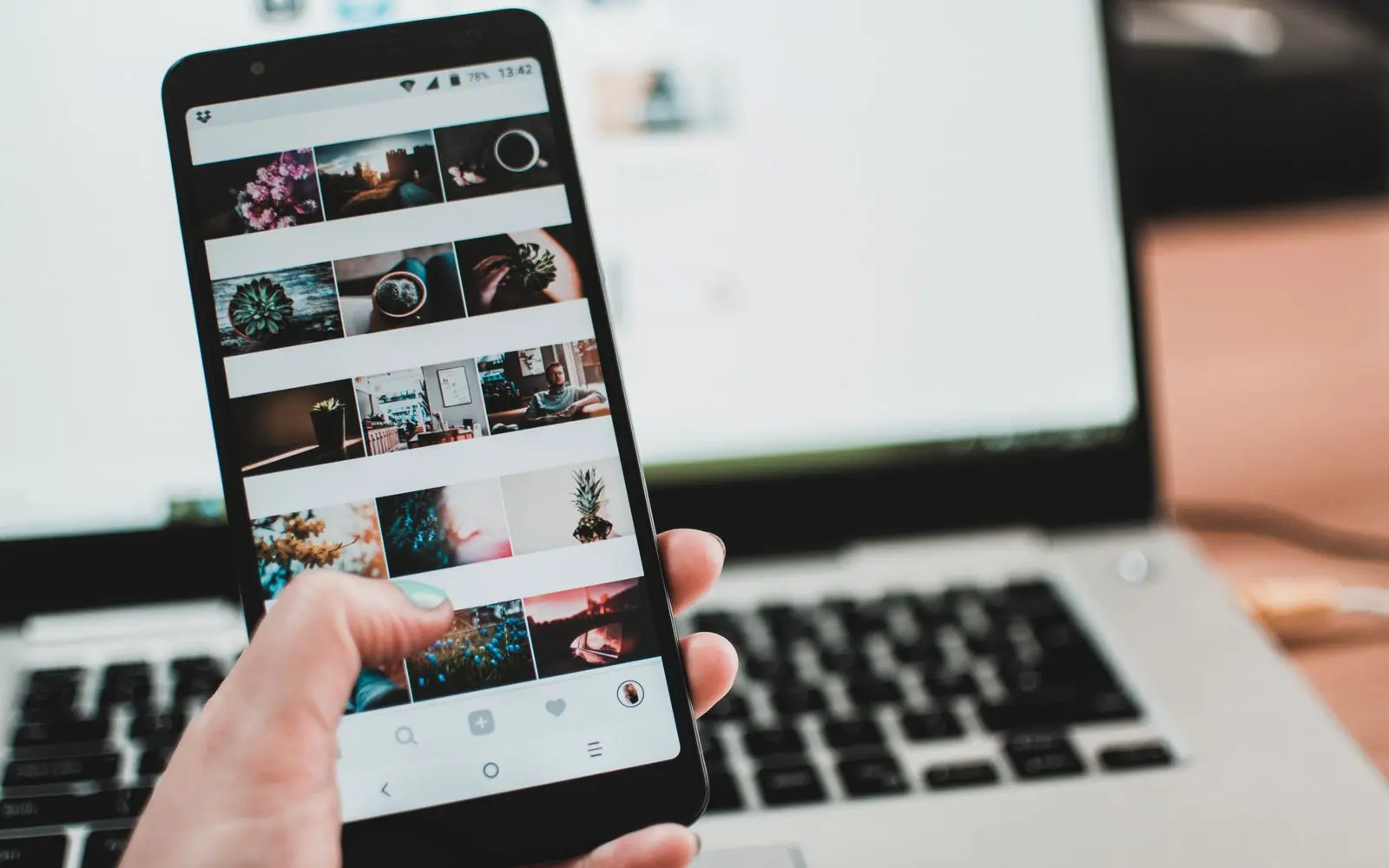 A hand scrolling a creator's grid of photos on a phone, in front of an open laptop.