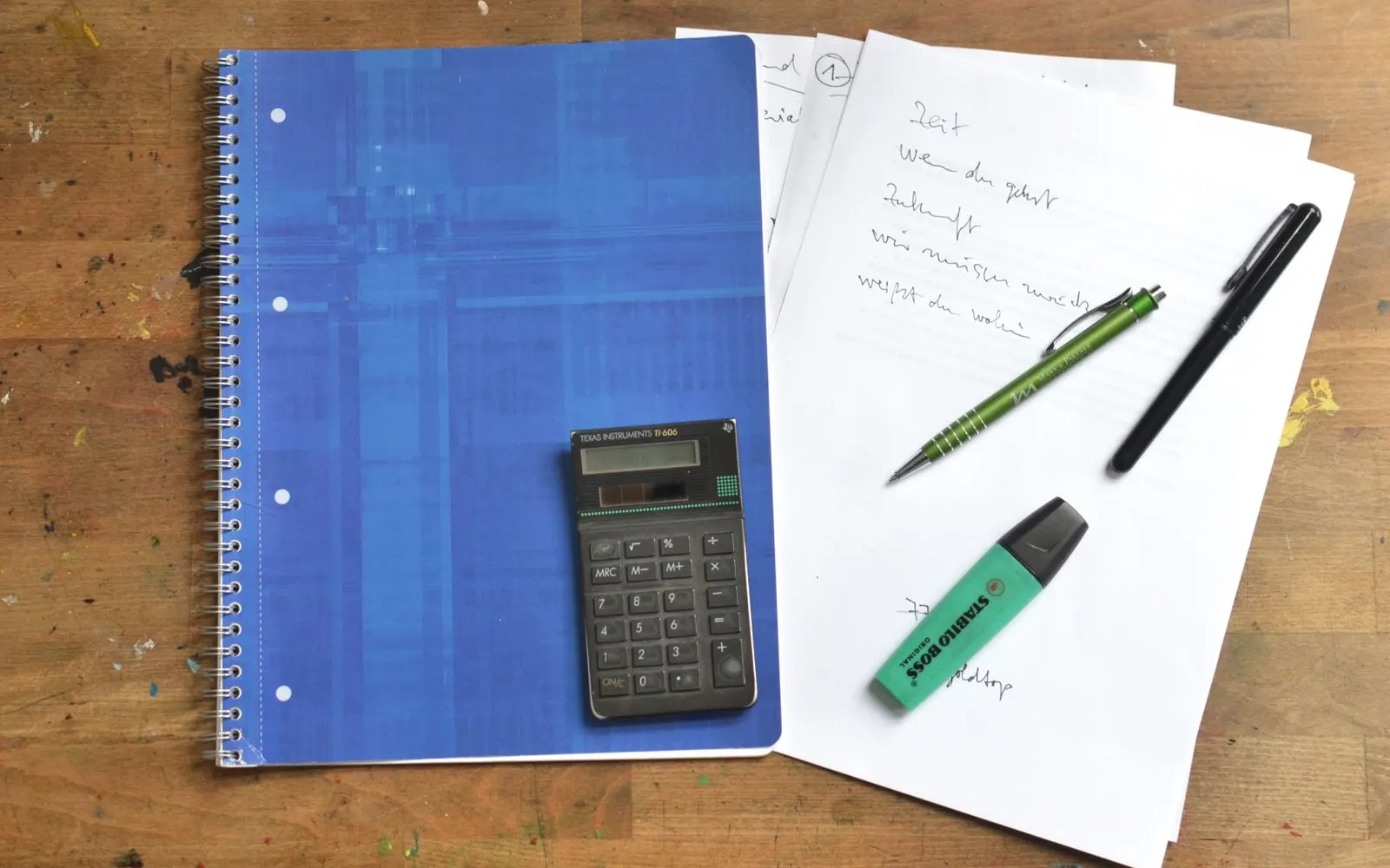 An open notebook of handwritten notes beside pens and a calculator on a wooden desk.